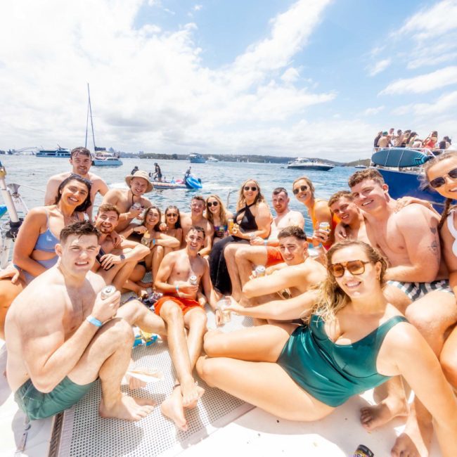 A group of people in swimsuits are sitting and standing closely together on a boat on a sunny day, with sailboats and blue skies visible in the background. They're enjoying a Catamaran party Sydney style.