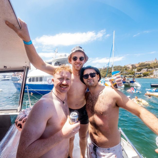Three men in swim trunks and sunglasses stand together on a boat, holding drinks, with a lively waterfront scene and people floating on inflatables in the background during a luxury yacht hire Sydney.