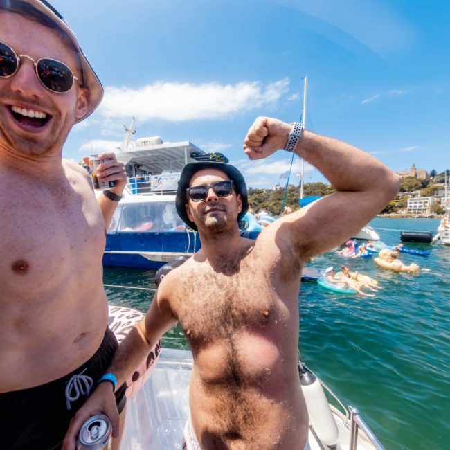 Two men posing shirtless on a boat, one flexing his bicep and the other smiling widely, both sporting sunglasses. Boats and people in the water are visible in the background. Enjoying a private yacht charter Sydney Harbour, they embody the joy of being out on the water.