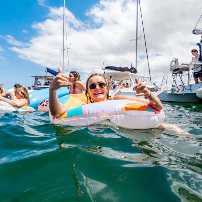A person in sunglasses floats on an inflatable ring in the sea, surrounded by others on inflatables and nearby boats, including a lively catamaran party Sydney is known for, all enjoying a sunny day.