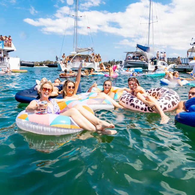 A group of people float on colorful inflatable rafts in a body of water near boats and other people. The weather is sunny with a partly cloudy sky, perfect for a Sydney boat party hire.