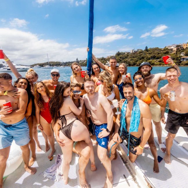 A group of people in swimwear holding red cups, posing on a catamaran with a scenic waterfront background, enjoying a Sydney boat party hire.