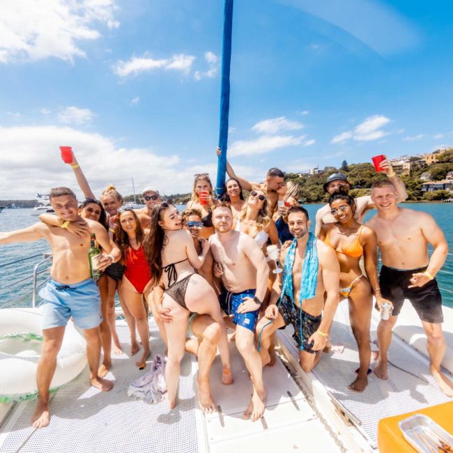 A group of people in swimwear enjoying a sunny day on a luxury yacht hire in Sydney, smiling and raising red cups, with a coastal backdrop in the distance.