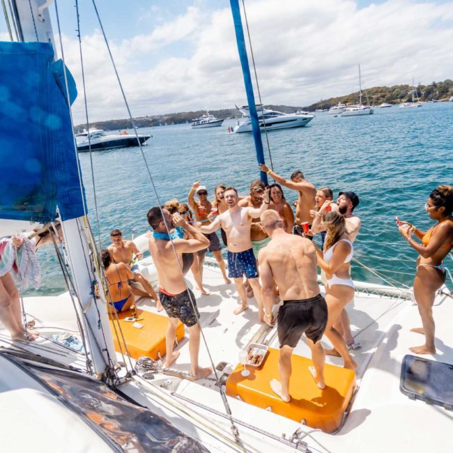 A group of people are having a lively gathering on a sailboat in a sunny and vibrant setting, surrounded by other boats on the water. Some are posing and taking pictures, enjoying what appears to be an exciting Sydney boat party hire.