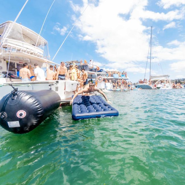 People gather on and around boats docked close to each other on a sunny day, with some standing on inflatable objects in the water. Enjoy a Sydney boat party hire or consider luxury yacht hire Sydney for an unforgettable experience.