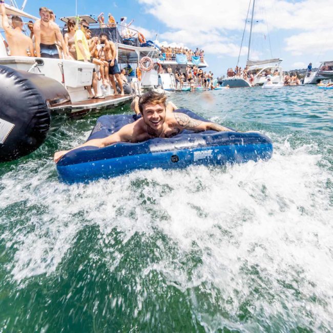 A man rides an inflatable mattress in a crowded area with boats and people swimming on a sunny day, enjoying the lively atmosphere of a catamaran party Sydney.