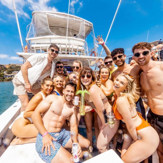 A group of people in swimwear pose for a photo on a boat under a sunny sky. Some are holding drinks and smiling at the camera. The water and coastline create the perfect backdrop for this fun-filled Sydney boat party hire.