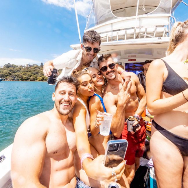 A group of people in swimwear are on a luxury yacht hire in Sydney, capturing a selfie. The background features sparkling water, lush trees, and other boats. One person holds a drink in a novelty cup shaped like a palm tree.