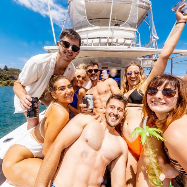 A group of people in swimwear smile and pose for the camera on a boat under a sunny sky, holding drinks and having a good time, enjoying a private yacht charter in Sydney Harbour.