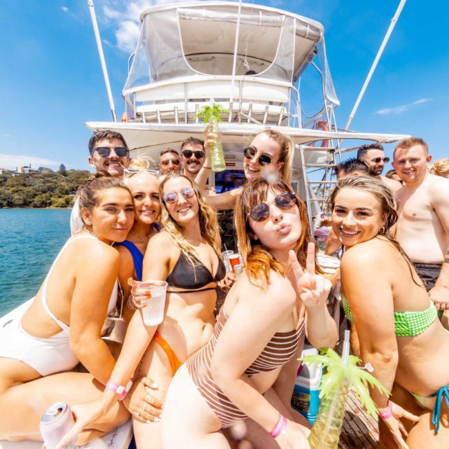 A group of people in swimwear posing on a boat under sunny weather with water and trees in the background. Some are holding drinks and smiling at the camera, enjoying a luxury yacht hire Sydney experience.