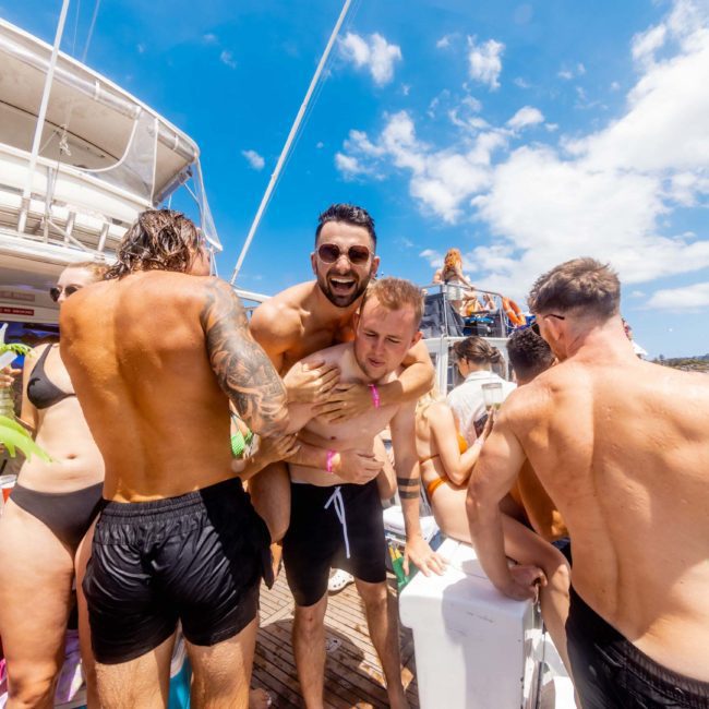 A group of people in swimwear are on a boat. Two men are lifting a man with a joyful expression. Other individuals are socializing or preparing to jump into the water, enjoying what could be one of the best Catamaran party Sydney experiences.