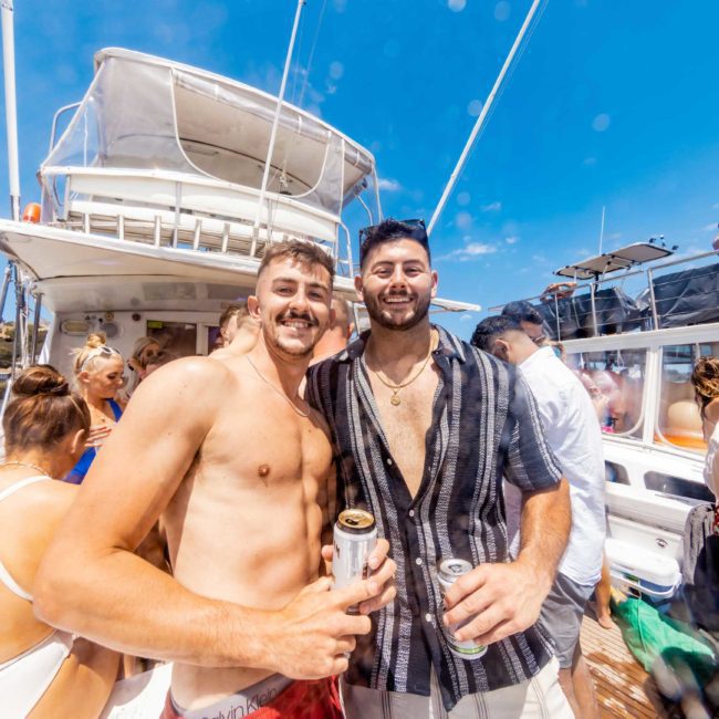 Two men pose smiling on a luxury yacht with drinks in hand, surrounded by other people enjoying a sunny day.
