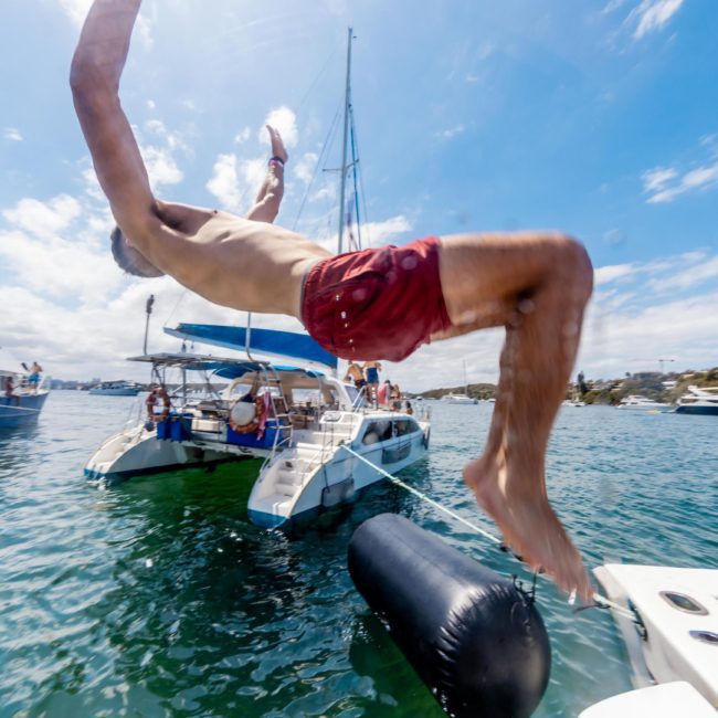 Person in red swim trunks mid-air as they jump into the water from a docked boat on a sunny day, with other boats and people in the background. Perfect scene for a Sydney boat party hire or private yacht charter on Sydney Harbour.