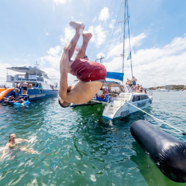 A person in red shorts is midair while doing a backflip off a boat into the water. Several boats and people are visible in the background, giving the scene the vibrant energy of a Sydney boat party hire.