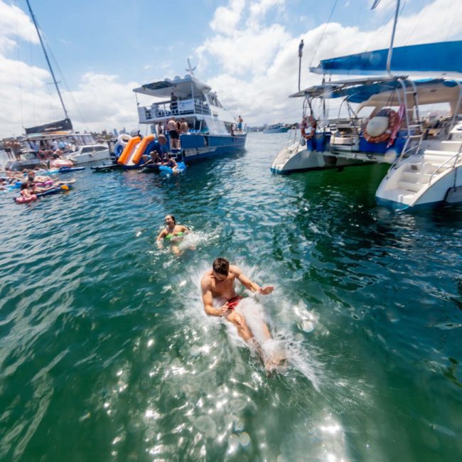 People swimming in water surrounded by anchored boats, with some individuals on inflatables and others on the boats. Sunny weather with a few clouds in the sky, perfect for a private yacht charter Sydney Harbour or even a lively Sydney boat party hire.