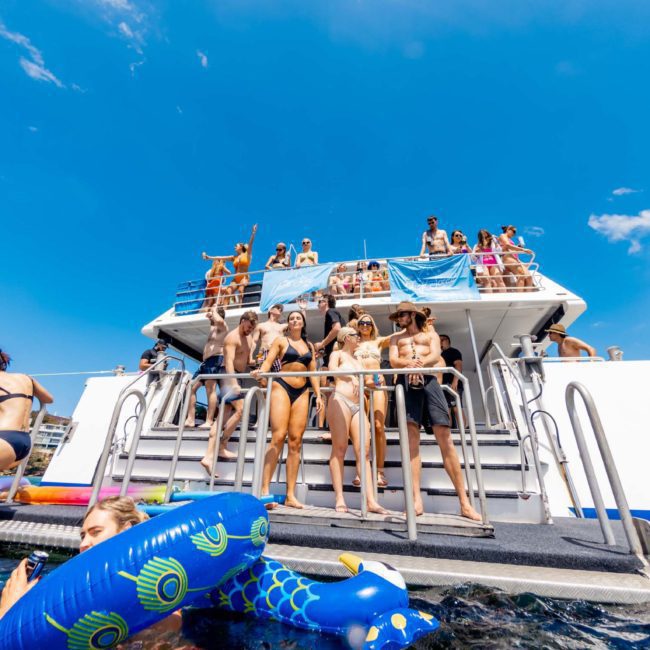 A group of people in swimwear stands on the back deck of a boat enjoying sunny weather. Some individuals are in the water with inflatable pool toys, making the most of their Luxury yacht hire Sydney.