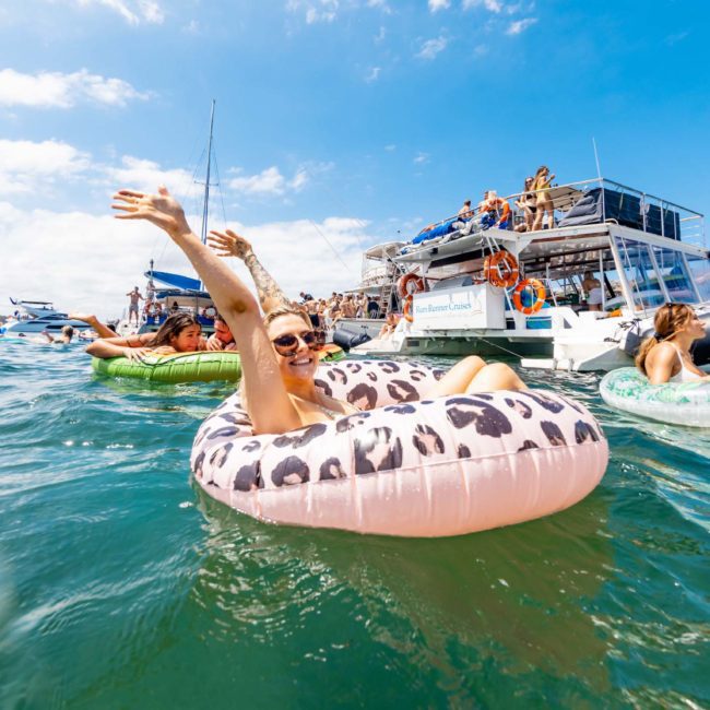 People enjoying a sunny day on the water, lounging in inflatable floats and swimming near boats, with several individuals visible on the boats in the background. This scene captures the essence of a lively Sydney boat party hire.