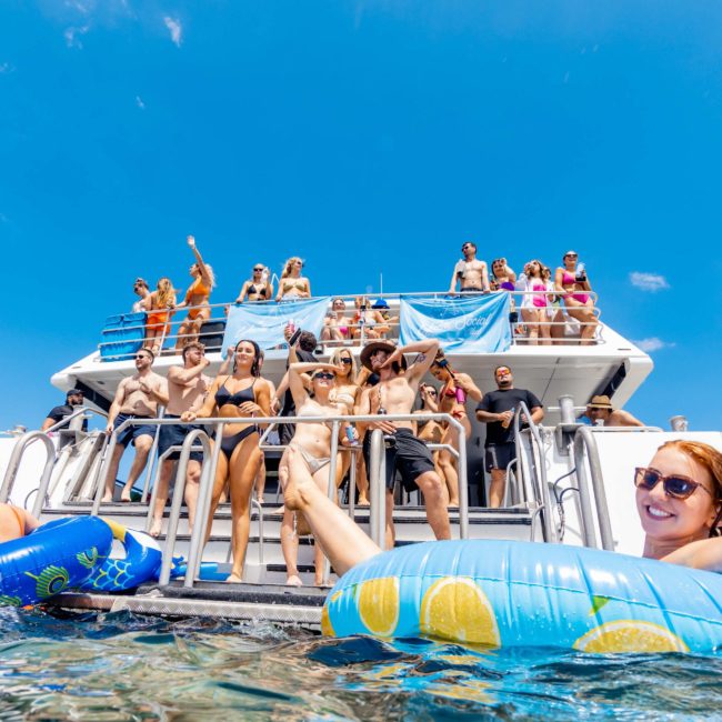A group of people in swimsuits stand and sit on a boat's boarding ladder, with some floating on inflatables in the water nearby, under a clear blue sky. Experience this perfect day with a private yacht charter in Sydney Harbour.