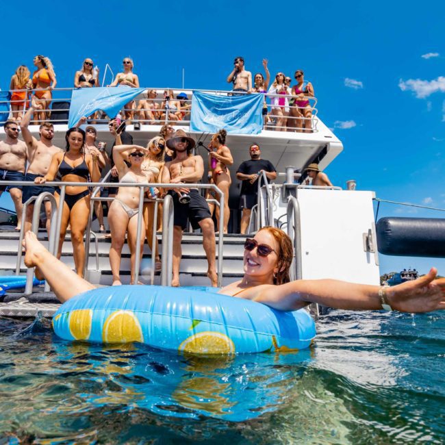 People on a Catamaran party in Sydney, some dancing and socializing, while one person floats on an inflatable ring in the water. The sky is clear and blue.