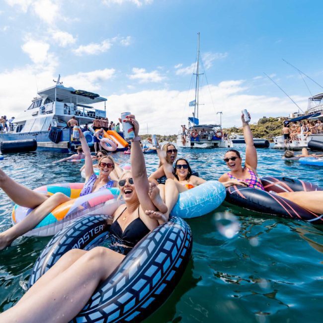 People in swimsuits are relaxing on inflatable rings in the water, raising their hands. Several boats are anchored nearby, including a luxury yacht hire Sydney option with a lively crowd aboard. The weather is sunny with a few clouds.