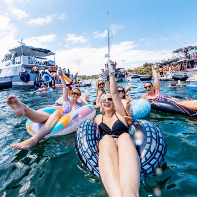 A group of people relax on inflatable pool floats in the ocean near several boats on a sunny day, enjoying the vibe of a Sydney boat party hire.