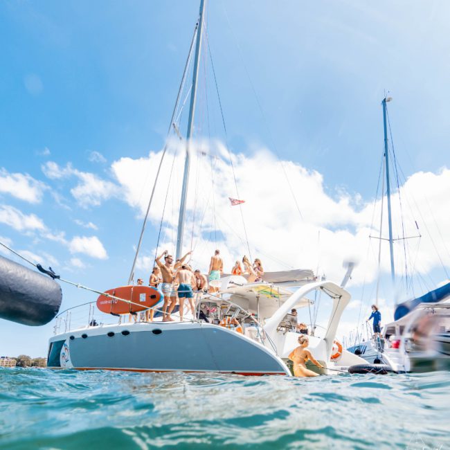 A group of people enjoys a sunny day on a luxury yacht hire in Sydney, with another sailboat nearby. Some are swimming, and the sky is clear with a few scattered clouds.