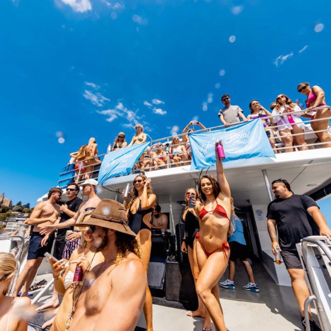 A group of people are gathered on a boat under a clear blue sky, some wearing swimsuits and sunglasses, enjoying a lively Sydney boat party hire.