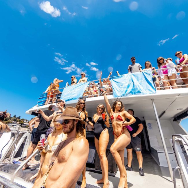 A group of people in swimwear enjoy a sunny day on a two-level boat, socializing and taking photos. The boat is surrounded by clear blue skies and scenic waters, perfect for a catamaran party in Sydney.