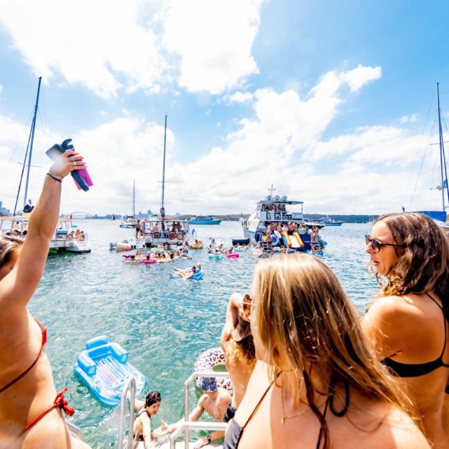 People on a boat enjoying a festive gathering of boats and swimmers in a sunny, blue water bay. Perfect for those seeking the vibrant experience of a catamaran party Sydney-style.