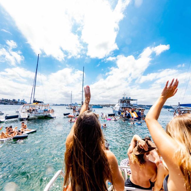 People enjoying a sunny day on boats and inflatable rafts in a lively harbor with clear blue skies, calm waters, and an atmosphere that feels just like a Sydney boat party hire.