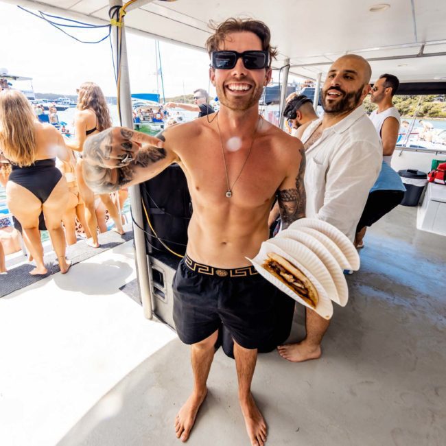 A man in black swim trunks smiles and holds a coiled hose on a luxury yacht filled with people in swimsuits. Another man stands behind him, and there are more people and a body of water in the background, enjoying what looks like an unforgettable Sydney boat party hire.