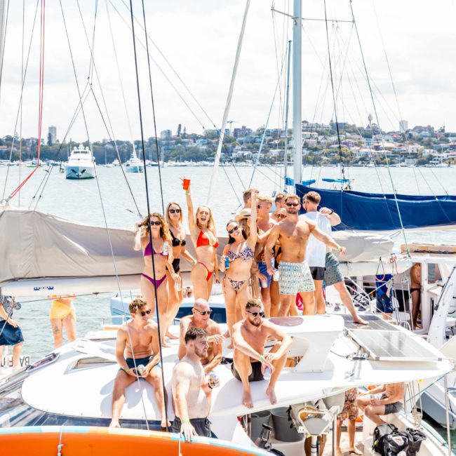 A group of people in swimwear stand and sit on a sailboat, raising their hands and drinks, with a cityscape in the background, enjoying their private yacht charter Sydney Harbour.