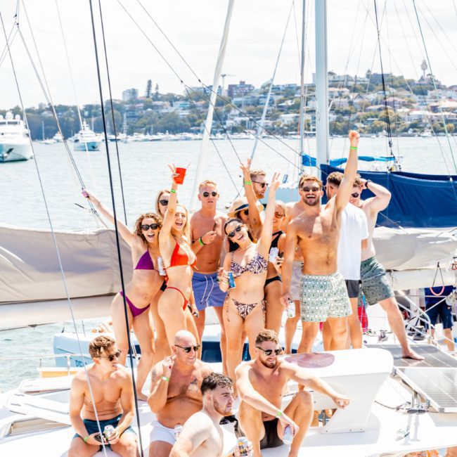 A group of people in swimwear are standing and sitting on a boat, smiling and raising their arms in celebration. The background features a body of water and houses on the shoreline, capturing the fun atmosphere of corporate boat events Sydney.