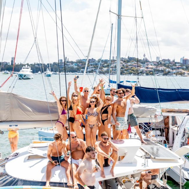 A group of people in swimsuits stand and pose on a luxury yacht hire Sydney in a sunny coastal area with other boats and a city skyline visible in the background.