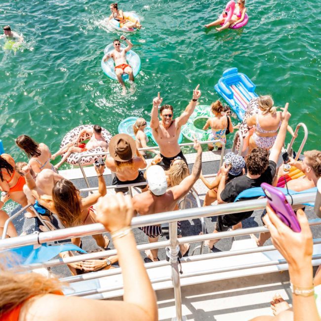 A group of people enjoying a sunny day on a private yacht charter in Sydney Harbour, swimming and floating on inflatable devices in the water. Some are on the yacht's deck, while others are in the water.