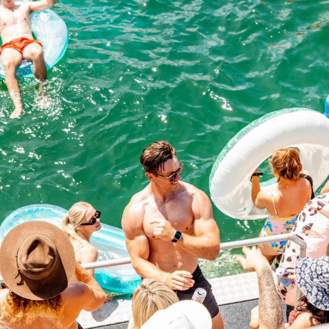A group of people enjoys a sunny day on a catamaran party in Sydney, with some in the water on inflatable floaties. One man flexes his muscles on the boat while others socialize and bask in the sun.