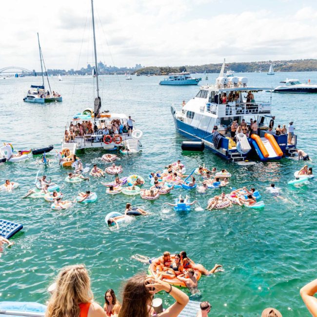People on boats and inflatable floats enjoying a sunny day on the water, with the city skyline visible in the background. In the mix, a luxury yacht hire Sydney adds an elegant touch to this perfect day.