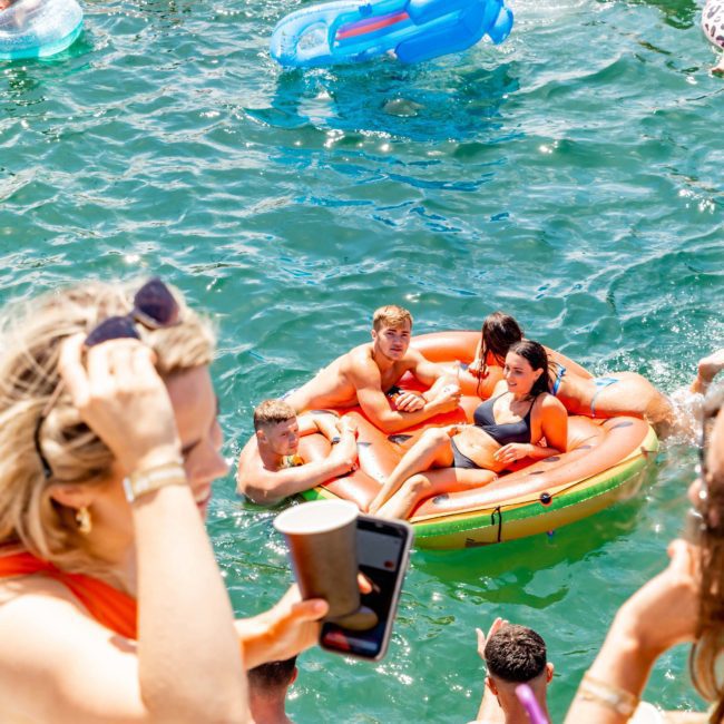 People relax in colorful floaties on a body of water, socializing and enjoying drinks in the sun. In the foreground, someone captures a moment with a drink and phone. Consider DJ boat hire Sydney for an unforgettable experience like this!