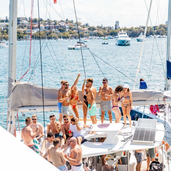 A group of people in swimwear are gathered on a boat, with some dancing on the deck. Other boats and a coastal town are in the background on a sunny day, enjoying a vibrant catamaran party Sydney style.
