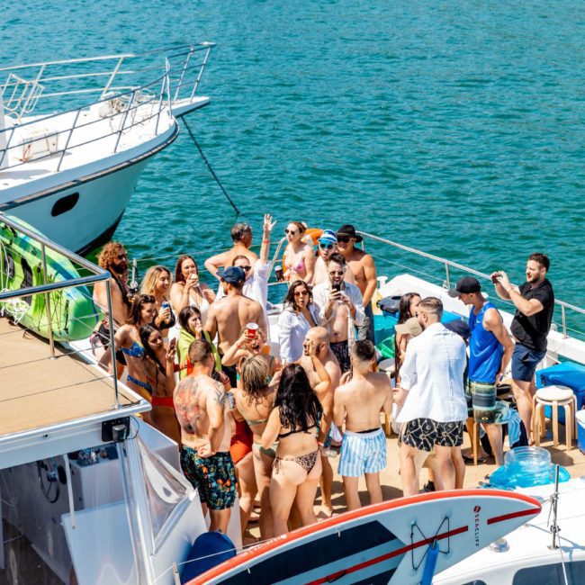 A group of people in swimwear enjoying a sunny day on a boat, gathered closely together, with another boat docked nearby on a clear blue water backdrop. Perfect for those looking into DJ boat hire Sydney for an unforgettable experience.