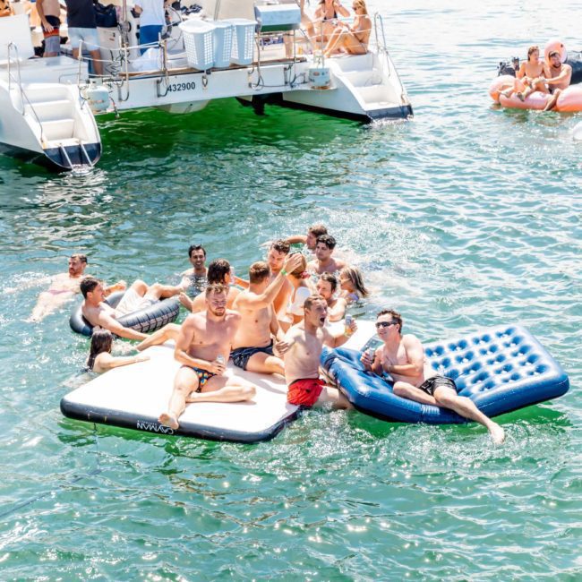A group of people are enjoying themselves on inflatable rafts in the water near a luxury yacht. Others are seen lounging on the boat and surrounding inflatables, making it a perfect Sydney boat party hire. The scene is lively and sunny.