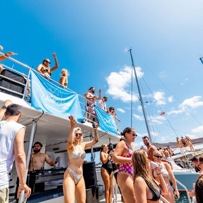 People in swimwear are dancing and socializing on a boat under a sunny sky, with banners displaying "Afloat Ibiza" hanging on the side of the boat. The scene echoes the vibrant atmosphere of a Catamaran party Sydney, capturing the essence of summer fun.
