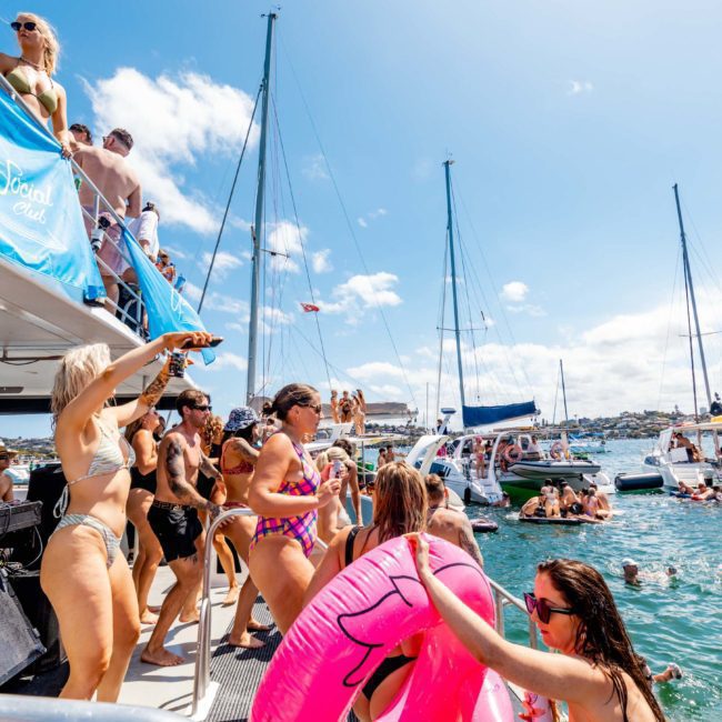 A group of people in swimwear are on and around a private yacht charter in Sydney Harbour, dancing and swimming with floating devices in the water. Several sailboats can be seen in the background under a sunny sky, creating the perfect setting for a Sydney boat party.