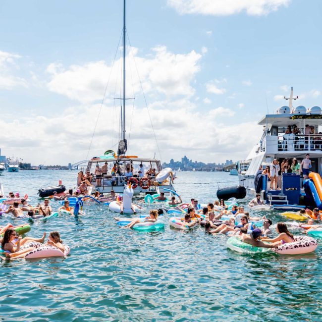 A large group of people in floaties enjoy a sunny day on the water near several boats, including one with slides, in a lively gathering. Consider corporate boat events Sydney for your next event to elevate the experience.