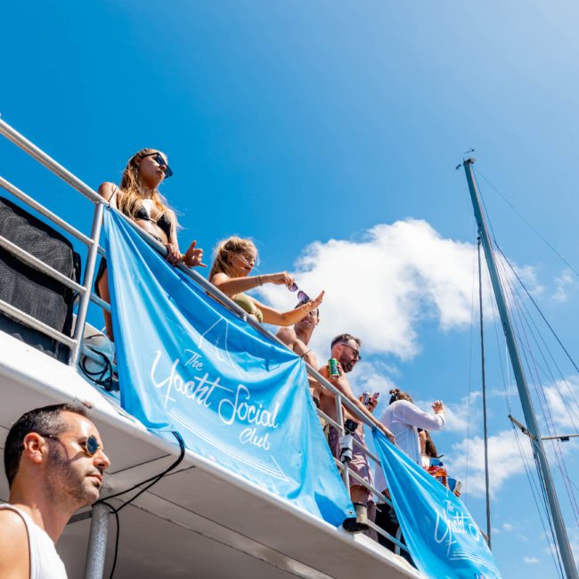 People are gathered on the deck of a private yacht charter in Sydney Harbour under clear blue skies, with a banner reading "The Yacht Social Club" displayed on the railing.