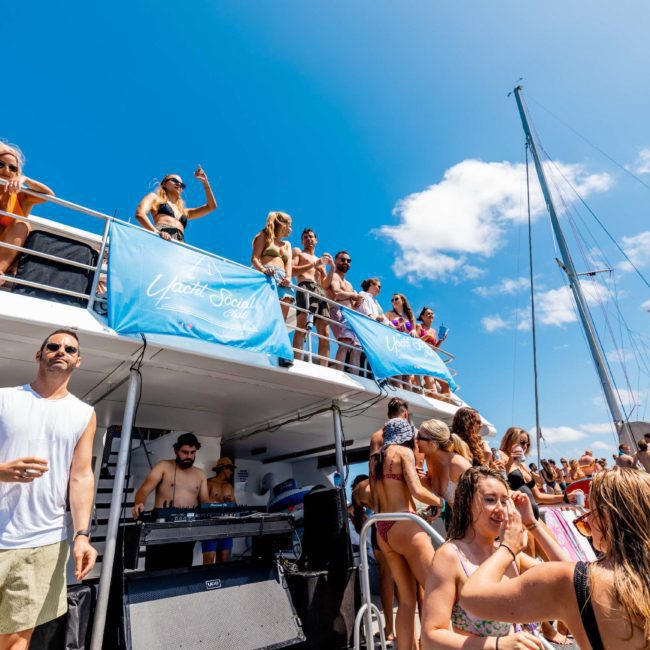 A large group of people, mostly in swimwear, are enjoying a sunny day on a double-deck boat with banners, a DJ, and clear blue skies in the background. This catamaran party in Sydney is perfect for soaking up the sun and having fun on the water with friends.