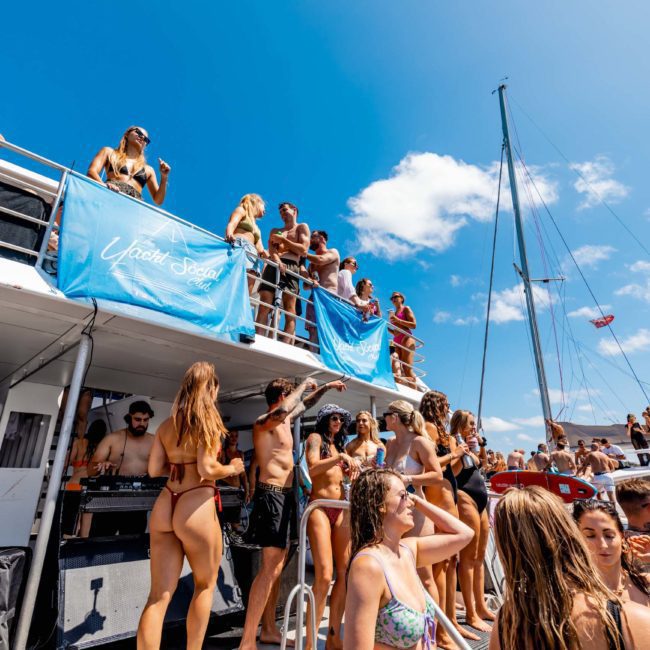 A large group of people, dressed in swimwear, are gathered on a luxury yacht for a Sydney boat party hire. Some are dancing while others are talking and drinking under a clear, sunny sky. A sailboat is visible in the background.