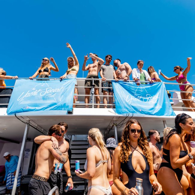 A large group of people in swimwear enjoy a sunny day on a two-level boat, with blue banners on the sides reading "Uplift & Social." Some people dance while others socialize and relax at this lively DJ boat hire Sydney event.