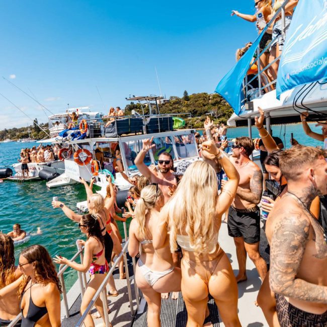 A group of people in swimwear are partying on luxury yachts in a sunny, tropical water setting with some dancing, swimming, and socializing.