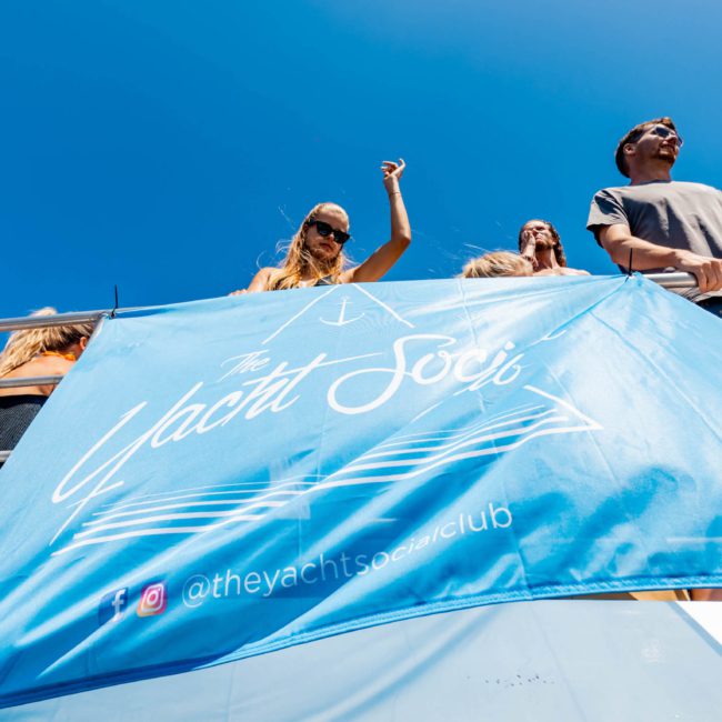 People on the upper deck of a yacht enjoying a sunny day. A blue banner with "The Yacht Soc" and social media handles hangs in the foreground, illustrating the perfect setting for a Corporate boat event Sydney or an unforgettable private yacht charter on Sydney Harbour.